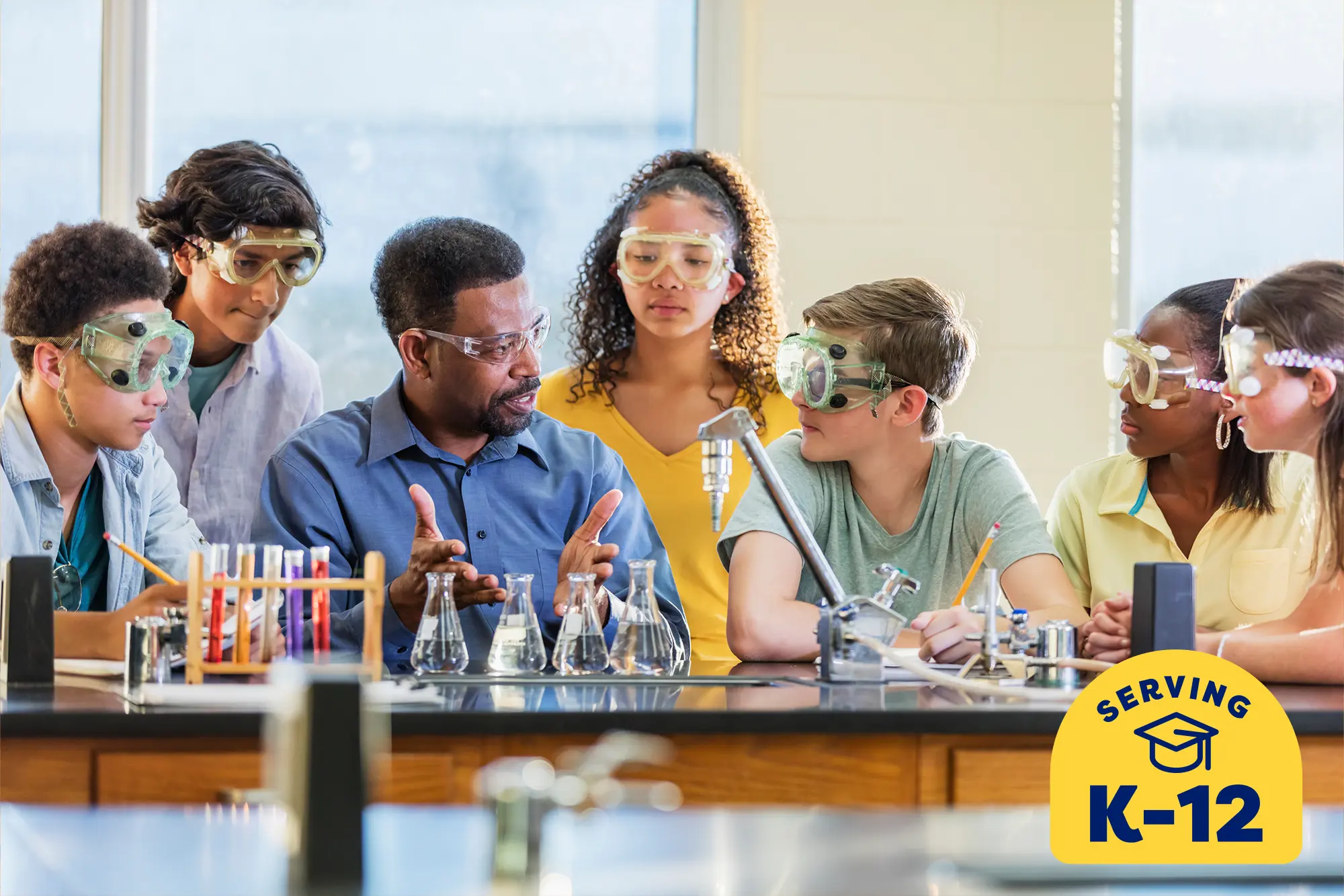 chemistry teacher sitting at a lab table explaining a project to several surrounding students