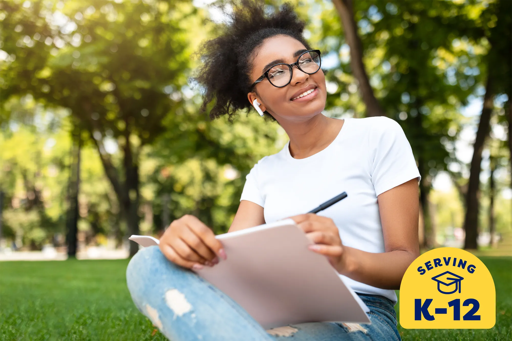 high school student sitting on the grass outside and looking up while writing in her notebook